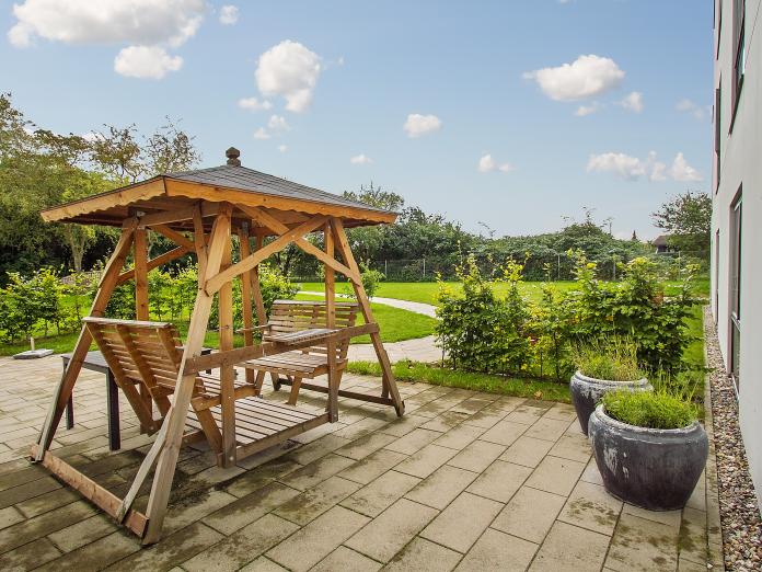 Fliseterrasse med hængesofa og grønne planter i krukker. Terrassen er omkranset af hæk og fra terrassen leder en sti ud til et grønt græsareal.