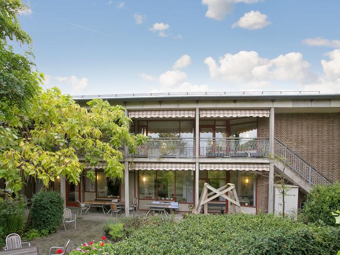 Bygningen med svalegang og markiser set fra gårdhaven, hvor der er store buske og høje træer samt borde og stole.