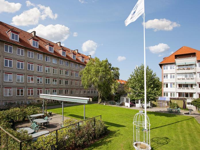 Terrasse med havemøbler og markise omkranset af rosenbuske. Terrassen ligger på en grøn græsplæne med flagstang, høje træer og espalier.