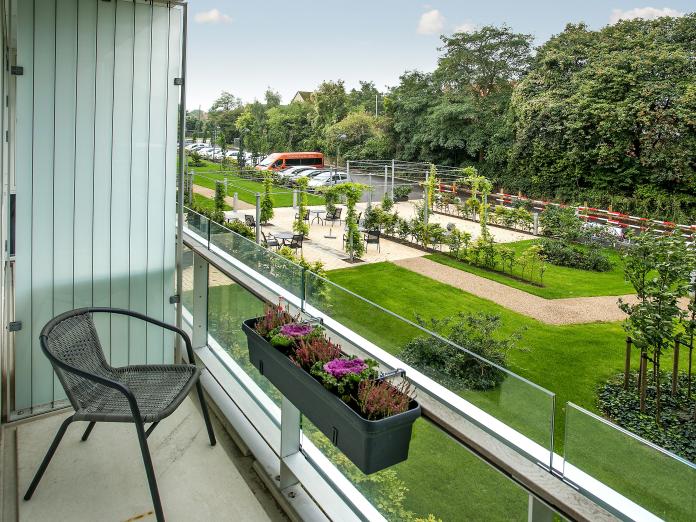 Altan med glasværn, stol og altankasser med blomster samt udsigt over park med græsplæner, gangstier, bede med stedsegrønt og træer.