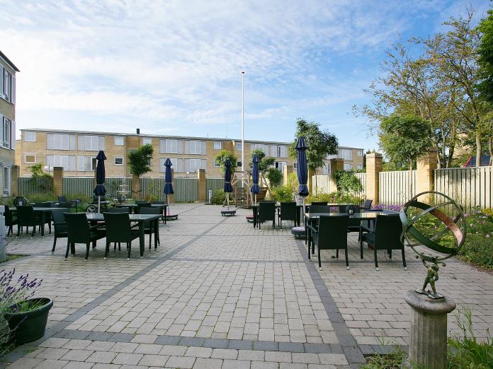 Stor fliseterrasse omkranset af hegn samt buske, bede og træer. Terrassen er indrettet med sorte havemøbler, parasoller og blomster i krukker.