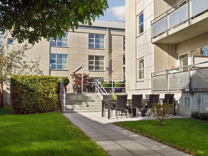 Mindre fliseterrasse med havemøbler. Omkring terrassen er græsplæner med bede, træer og buske.