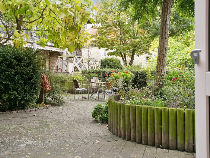 Lille krog af gårdhave med cafébord og stole omgivet af buske, højbede med forskellige blomster og høje træer. 