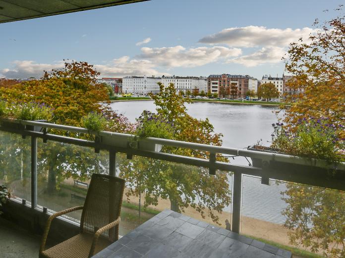 Udsyn fra altan ud mod store træer, grusstier og Sortedamssøen. Altanen er indrettet med bord, fletstol og altankasser med blomster.