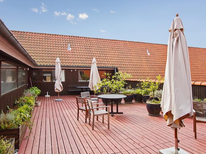 Stor træterrasse med højbede med grønne planter og blomster samt store krukker med grønne planter. Terrassen er desuden indrettet med havemøbler og parasoller. 