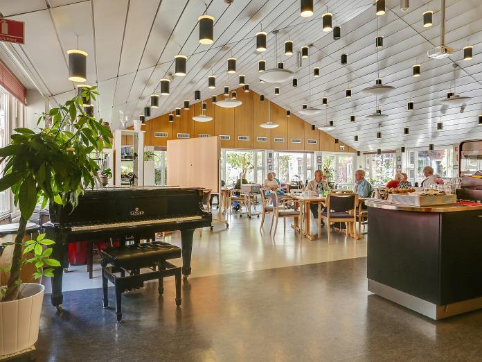 Fællesområde med borde, stole, flygel og grønne planter. 