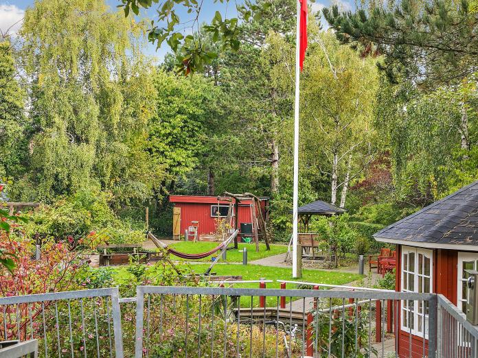 Grøn have med træer, buske, bed, græsplæne, parasoller, hængekøje, flagstang og et lille rødt tehus. 