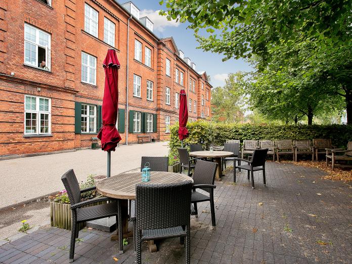 Fliseterrasse med kurvemøbler og runde borde samt parasoller og blomster i højbede.