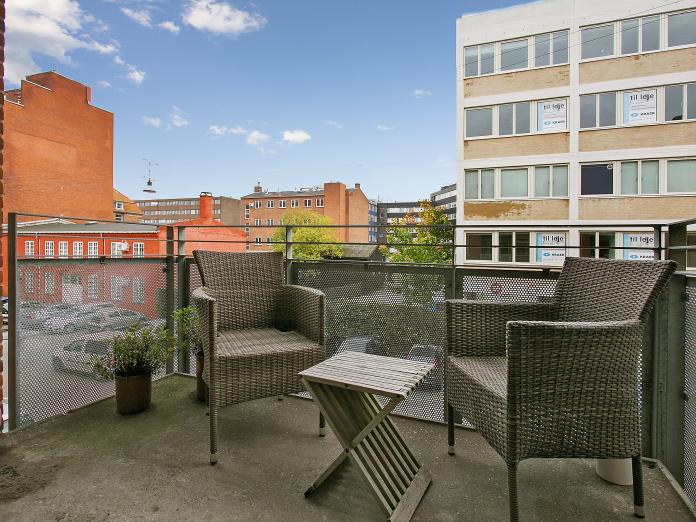 Privat altan med kurvemøbler og borde samt planter i krukker. Fra altanen ses nyere kontorbyggeri, ældre huse og lejlighedskomplekser samt grønne træer.