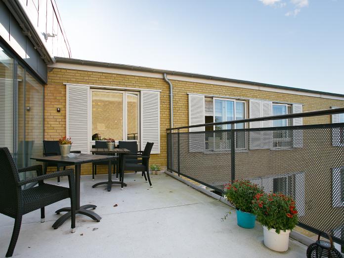 Åben, fælles altan med sorte havemøbler og blomster i krukker.