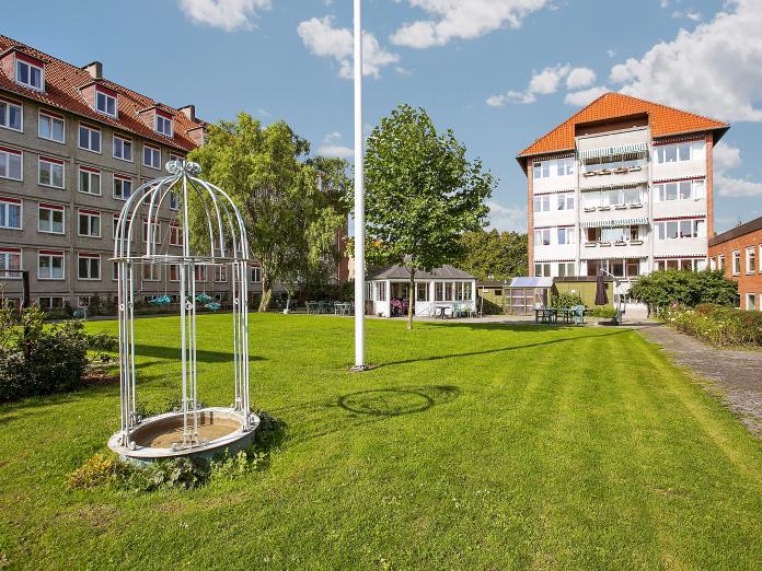 Grønt græs med bede, buske, espalier, flagstang og høje træer. I baggrunden ses en terrasse med udestue og drivhus.