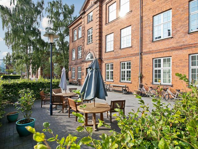 Flisegårdhave med havemøbler i mørkt træ, parasoller og grønne planter i krukker. Gårdhaven er omkranset af bøgehæk og i baggrunden ses høje piletræer.