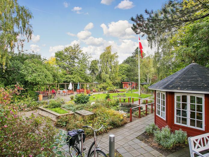 Grøn have med træer, buske, bed, græsplæne, parasoller, hængekøje, en cykel, flagstang og et lille rødt tehus. 