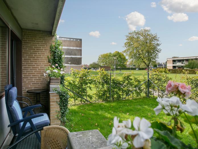 Lejlighed med lille terrasse møbleret med havemøbler og en lille have med blomster i blomsterkasser samt grønt græs omkranset af en hæk. 