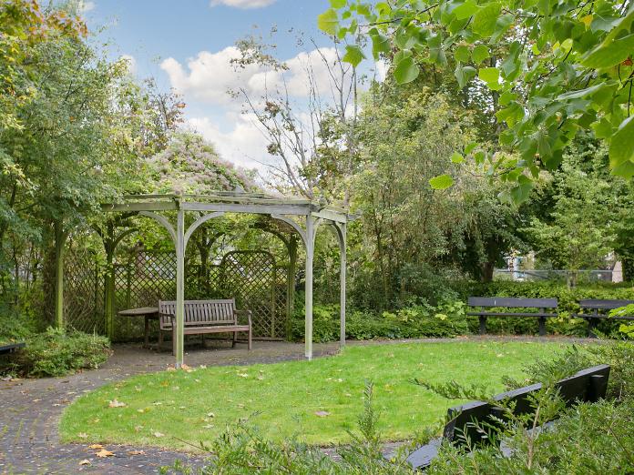 Frodig, grøn have med pavillon, græsplæne, bede og høje træer samt bænke og et bord.