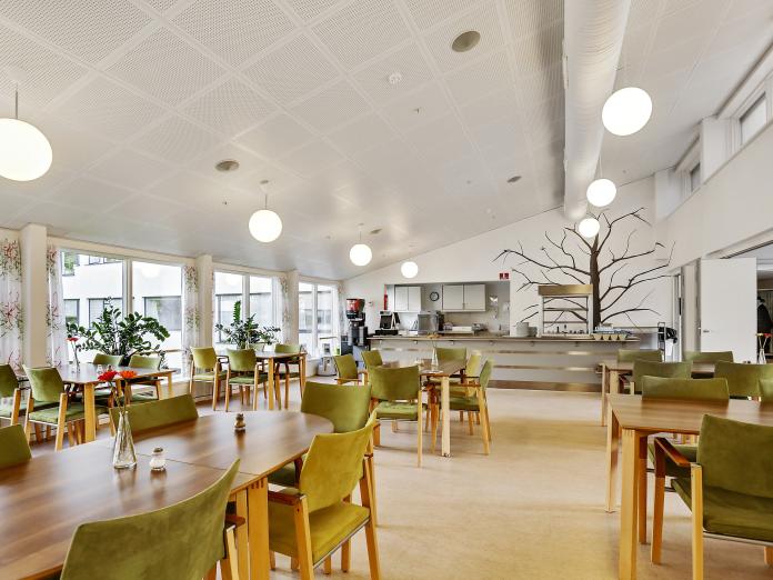 Fællesrum med 6 borde og grønne stole, højt til loftet og køkken i baggrunden. På væggen er malet et stort træ uden blade.