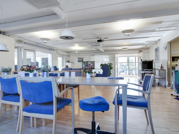 Stort fælles rum med køkken samt borde med stole omkring og et sofahjørne med fjernsyn.