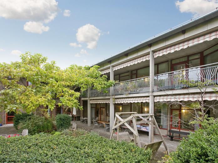 Bygning i røde sten og beton med svalegang og rød-og-hvidstribede markiser. Foran bygningen er trimmede buske, høje træer og spredte siddeområder.