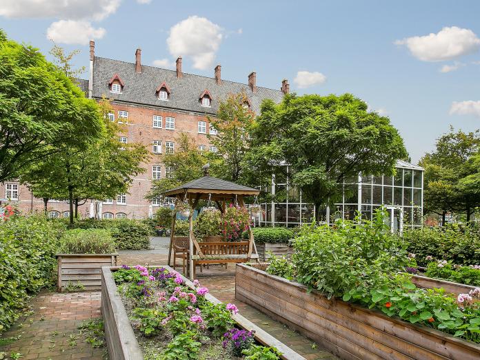 Gårdhave med flere højbede med blomster, krydderurter og grønt, buske og store træer samt en hængesofa og i baggrunden et stort drivhus.