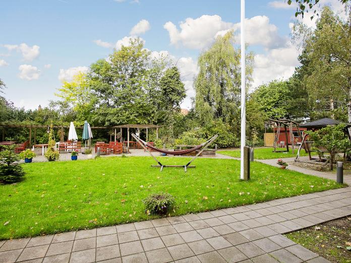 Græsplæne og stier, hængekøje, flagstang og en terrasse med parasoller.