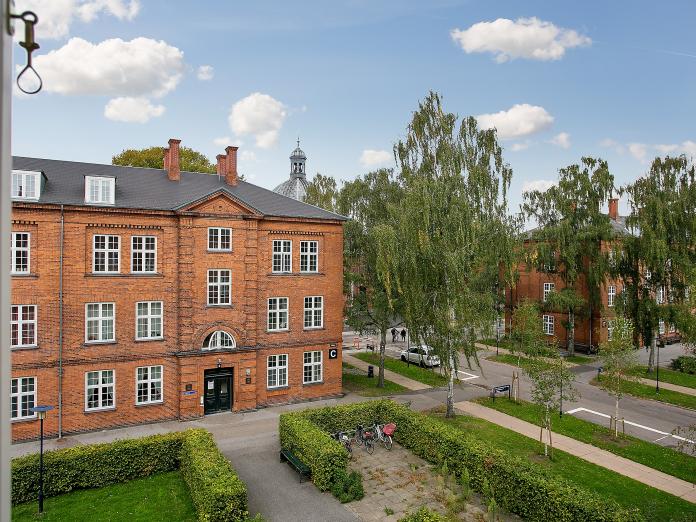 Ældre, rødt murstenbyggeri med hvide vinduer og sort skifertag. Foran bygningen er der høje birketræer og gårdhaver omkranset af grønne buske.