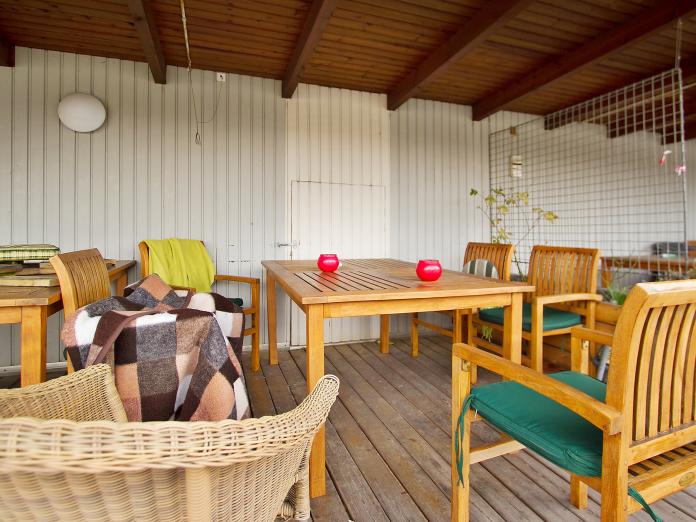 Overdækket terrasse med træhavemøbler, tæpper og grønne planter.