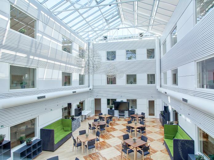 Stor, lys atriumgård med glastag. Rummet er indrettet med akustiksofaer og reoler langs væggene samt mange mindre borde med stole omkring.