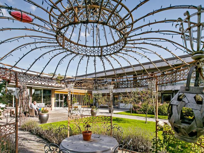 Have med flisegange og terrasser med havemøbler og bænke. I forgrunden en pergola i jern med lyskæder. Omkring pergolaen er der plantet lavendel og en rosenbusk samt grønt græs. 