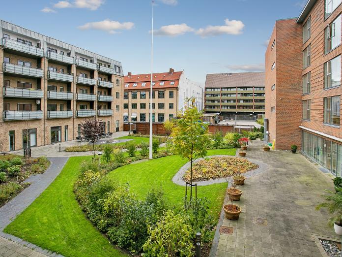 Gårdhave med græsplæne, bede med stedsegrønne buske og træer samt gangstier mellem terrasser, hvor der står blomster i krukker. 