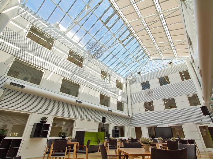 Stor, lys atriumgård med glastag. Rummet er indrettet med akustiksofaer og reoler langs væggene samt mange mindre borde med stole omkring.