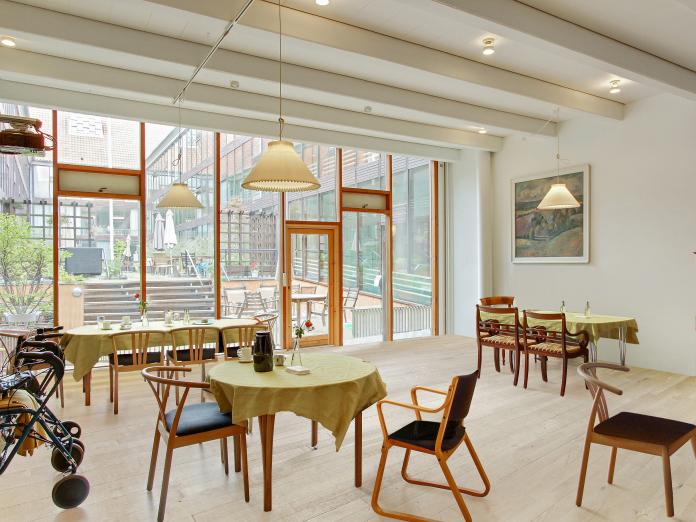 Lyst rum med glasfacade ud mod atriumgård. Rummet er møbleret med borde med dug på og med stole omkring bordene.