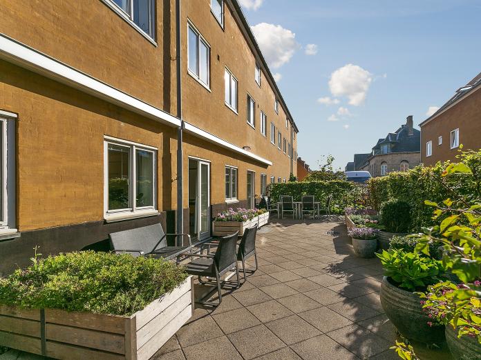 Fælles fliseterrasse omkranset af hæk. Terrassen er indrettet med blomsterkasser, blomster i krukker og havemøbler.