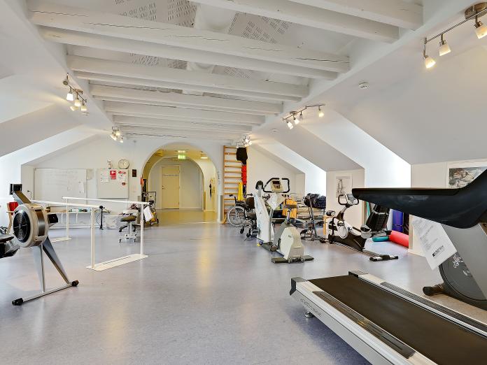 Lyst motionsrum med fritlagte bjælker. Rummet er indrettet med flere træningsmaskiner som løbebånd og kondicykel samt en gangbarre og en ribbe.