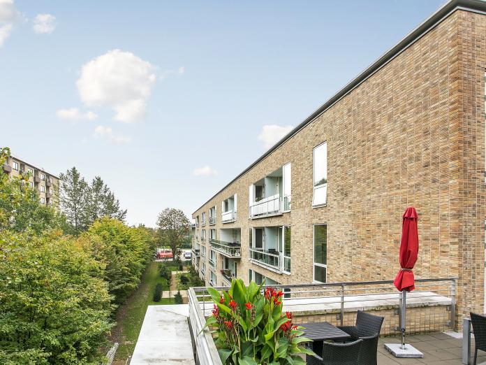 Kig langs facaden af moderne bygning i lyse sten med lukkede altaner. Foran bygningen er grønt græsareal med store træer. 