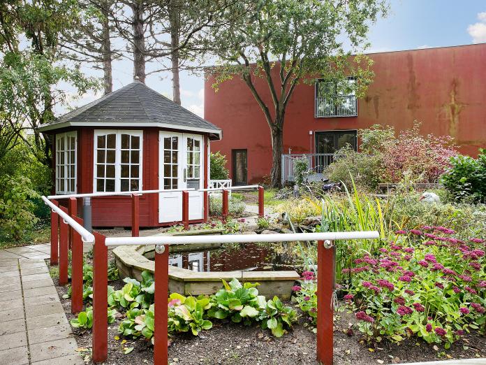 Et rødt tehus, et bed, en lille dam og store træer i baggrunden.