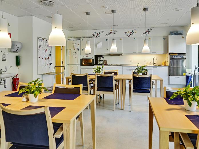 Fælles spiseområde indrettet med borde og stole samt med køkken i den ene ende af rummet. Køkkenet består af ovn, køleskab, vask og opvaskemaskine.