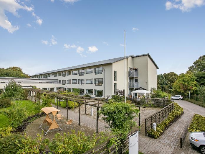 Bispebjerghjemmet set udefra med vinduer, hvid-grå facade, indkørsel, terrasser, buske og træer fra Lersøparken i baggrunden. 