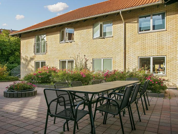 Gul murstensbygning med rødt tag. Foran bygningen er en gårdhave med blomsterbede og havemøbler.