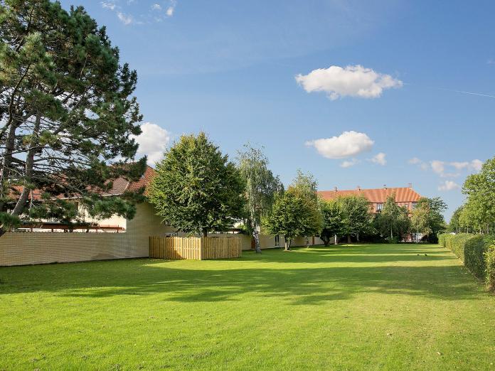 Aflang, grøn nyslået græsplæne med store, grønne træer omkranset af en gul murstensvæg og grønne hække. 