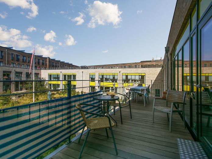 Stor fællesaltan med borde og stole. Fra altanen er der udsigt til de omkringliggende bygninger samt en stor grøn gårdhave.