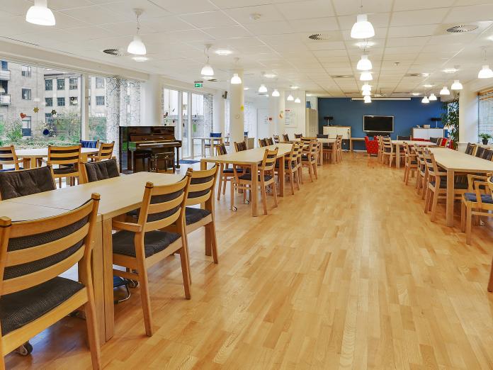 Stort fællesrum med langborde og stole, et klaver og gul til loft-vinduer ud til fælles gårdhave. Endevæggen er malet blå, mens de resterende vægge og lofter er malet hvide. 