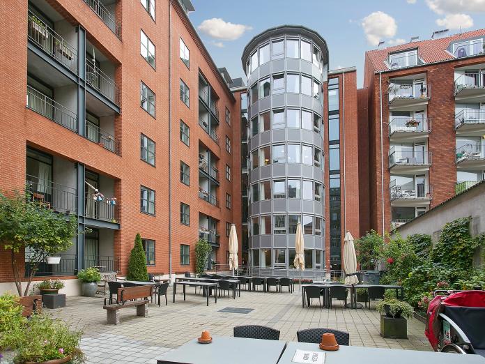 Moderne seksetagers bygning med altaner og en gårdhave foran. Gårdhaven er møbleret med borde, stole, parasoller og bænke samt bede, buske, træer og blomster i krukker.