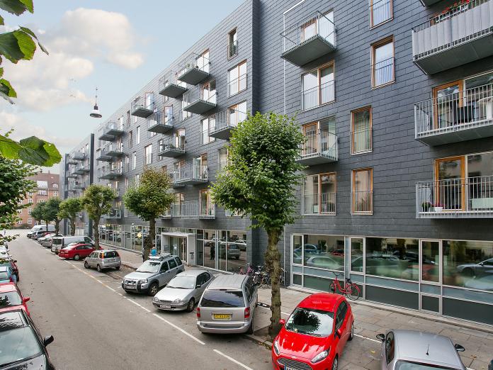 Moderne bygning set fra gaden. Bygningen er i mørkegrå sten, træfarvede og hvide vinduer samt stålaltaner. Foran bygningen er der parkeringspladser og høje, gamle træer.