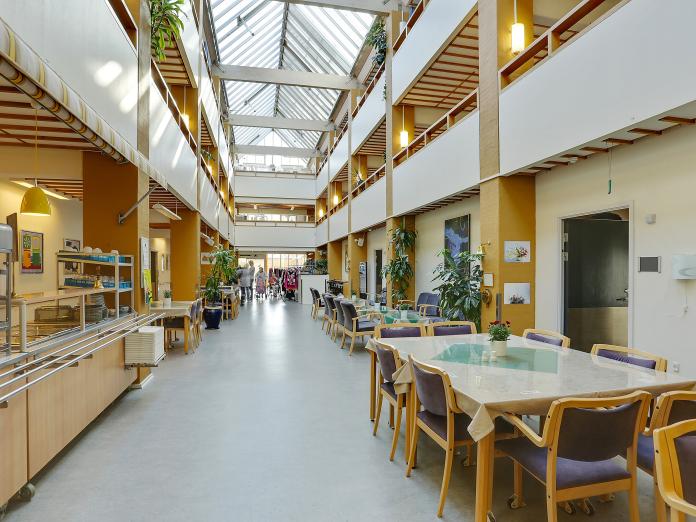 Store fællesrum i indendørs atriumgård. Rummet er indrettet med grønne planter, borde og stole samt et kantineområde.