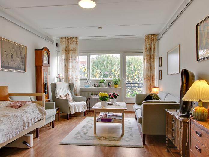 Lyst værelse med fransk altan. Værelset er indrettet med plejeseng og loftslift samt personlige møbler som sofa, lænestol, sofabord, kommoder og et bornholmerur.