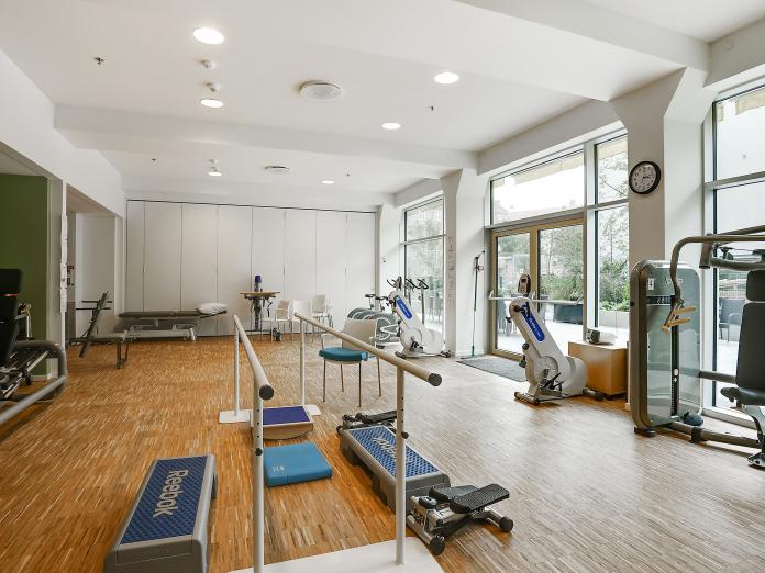 Motionsrum med store vinduespartier ud til gårdhave. Rummet indeholder mange maskiner samt gangbarre og behandlingsbriks. 