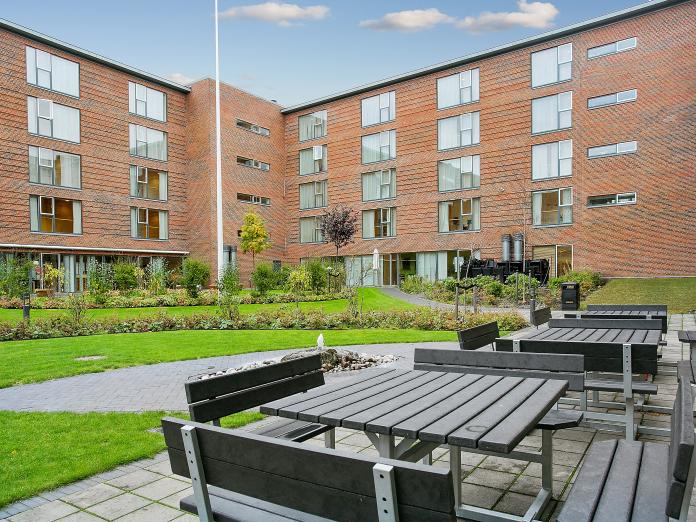 Moderne bygning i røde sten med græsplæne, bede og gangstier samt bord- og bænkesæt foran. 