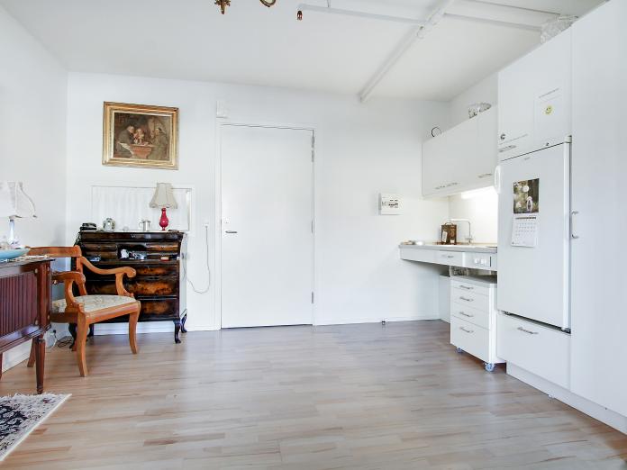 Lys stue med tekøkken og personlige møbler som skrivechatol, stol og billeder på væggen.