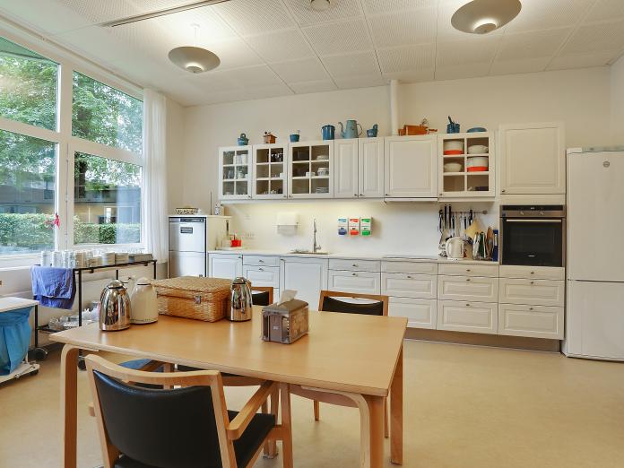 Stort fællesrum med køkken ved den ene væg samt bord med stole omkring.