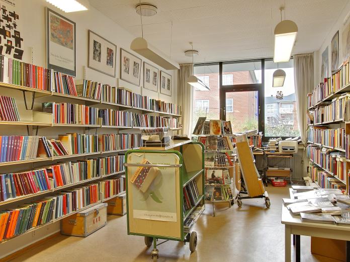 Bibliotek med reoler langs væggene fuld af bøger samt et bogstativ og en rullevogn med bøger. På væggene er billeder.